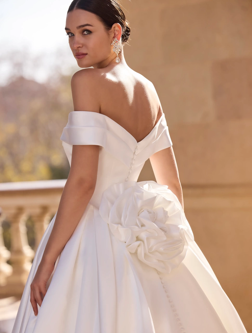Abito da sposa semplice da principessa con vita basca, scollo off-shoulder con dettaglio particolare, gonna strutturata ampia in mikado drappeggiata sui fianchi con fiore 3D scenografico sul retro