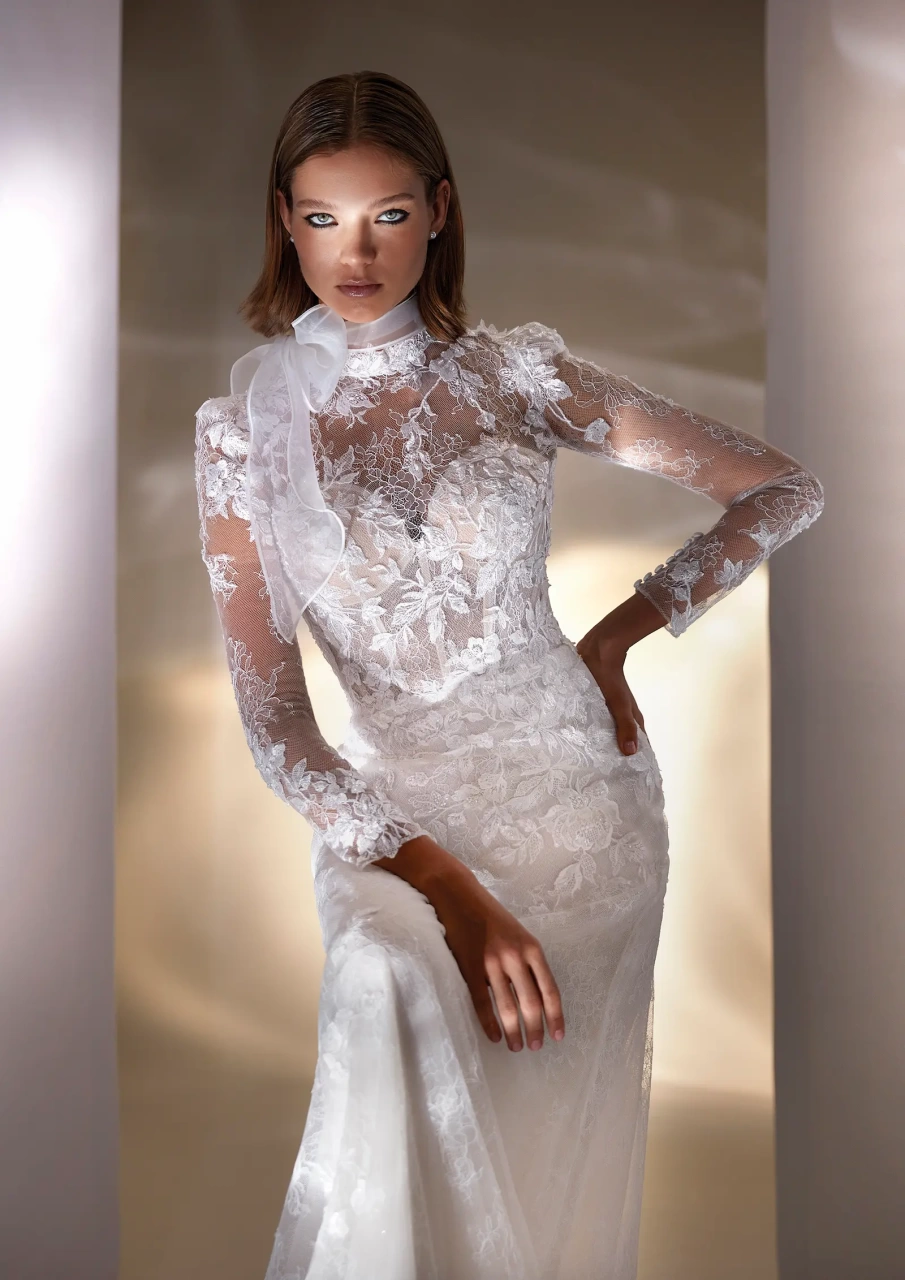 Abito da sposa con maniche lunghe in pizzo, collo alto con fiocco in tulle, corpino steccato, gonna scivolata aderente in tulle con applicazioni di pizzo floreale | Lo Specchio Atelier Abiti da Sposa a Verona
