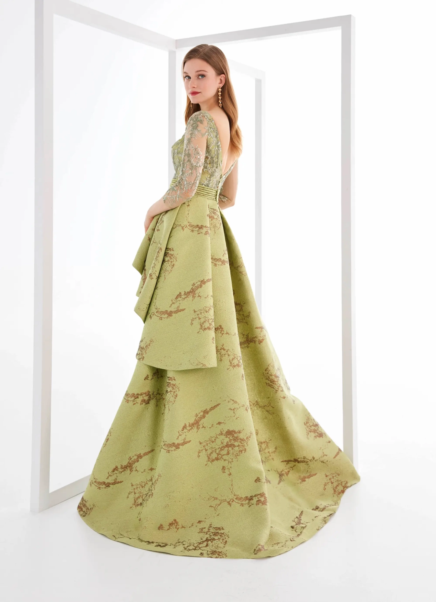 Vestito da cerimonia color verde chiaro lungo con maniche lunghe velate e pizzo ton-sur-ton, gonna lunga a sirena, sovragonna con elegante stampa a contrasto e pieghe asimmetriche sul fianco sinistro, bustino con scollo a V profondo velato - Lo Specchio Atelier Abiti da Cerimonia a Verona