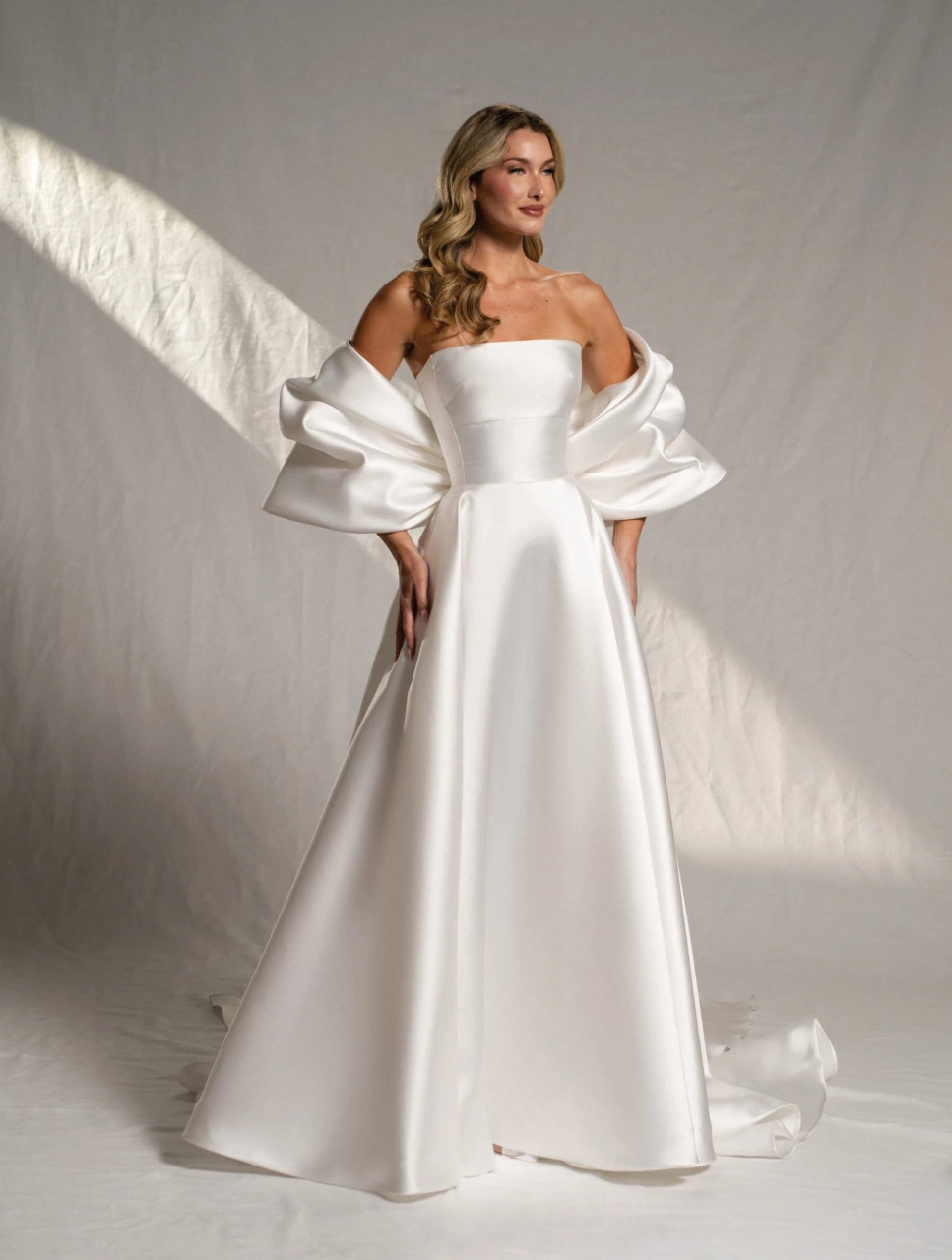 Abito da sposa semplice A-line in mikado lucido, scollo dritto senza spalline, dettaglio particolare di fiocco grande sulla schiena che funge da maniche grandi e voluminose