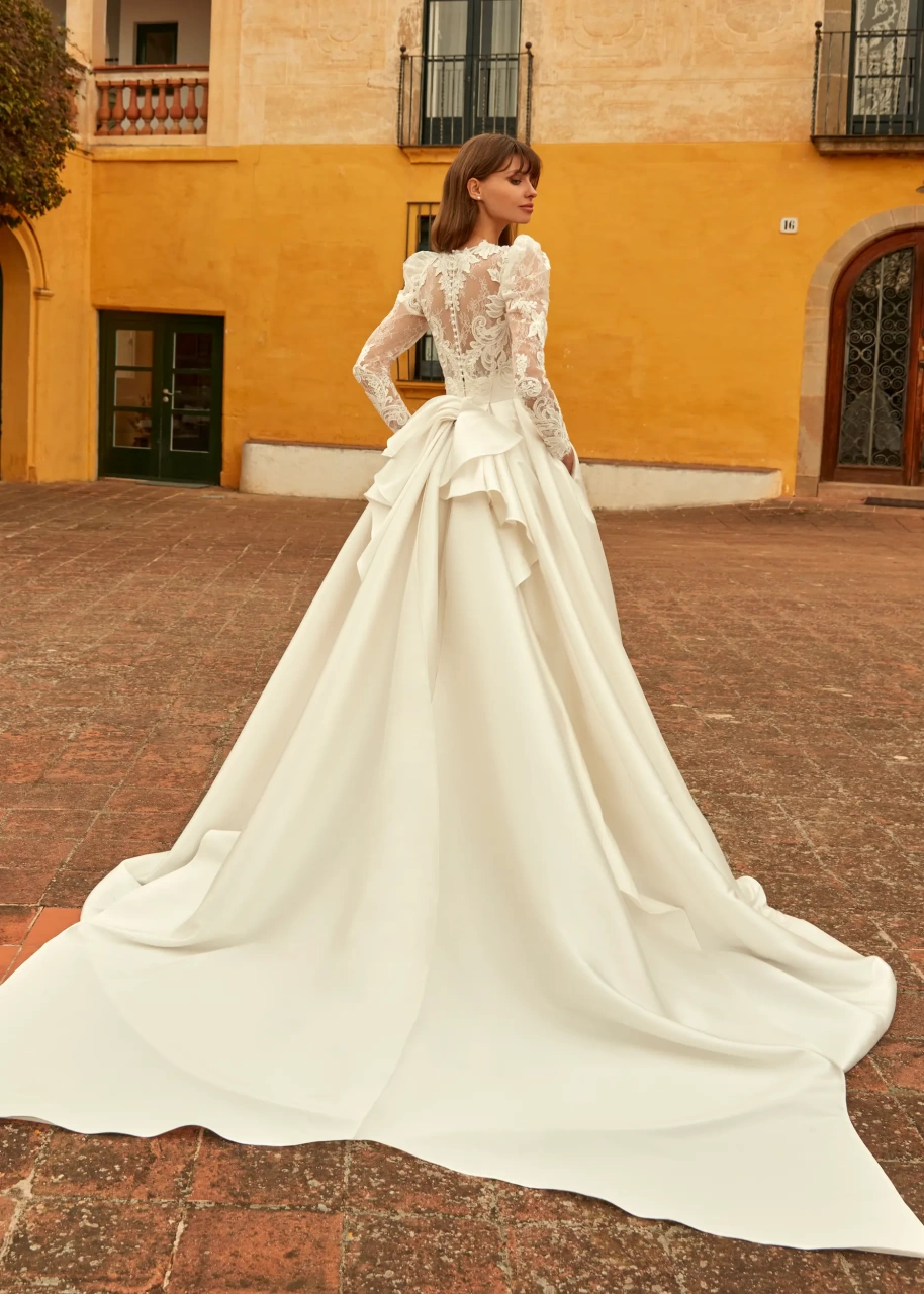 Abito da sposa particolare con maniche lunghe a sbuffo in pizzo effetto tattoo, scollo rotondo, gonna ampia A-line drappeggiata in mikado con fiocco drappeggiato sulla schiena e strascico lungo