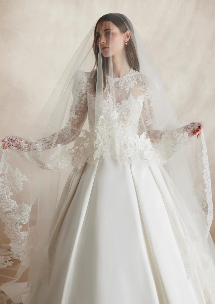Abito da sposa particolare con maniche lunghe a sbuffo in pizzo effetto tattoo, scollo rotondo, gonna ampia A-line drappeggiata in mikado con fiocco drappeggiato sulla schiena e strascico lungo