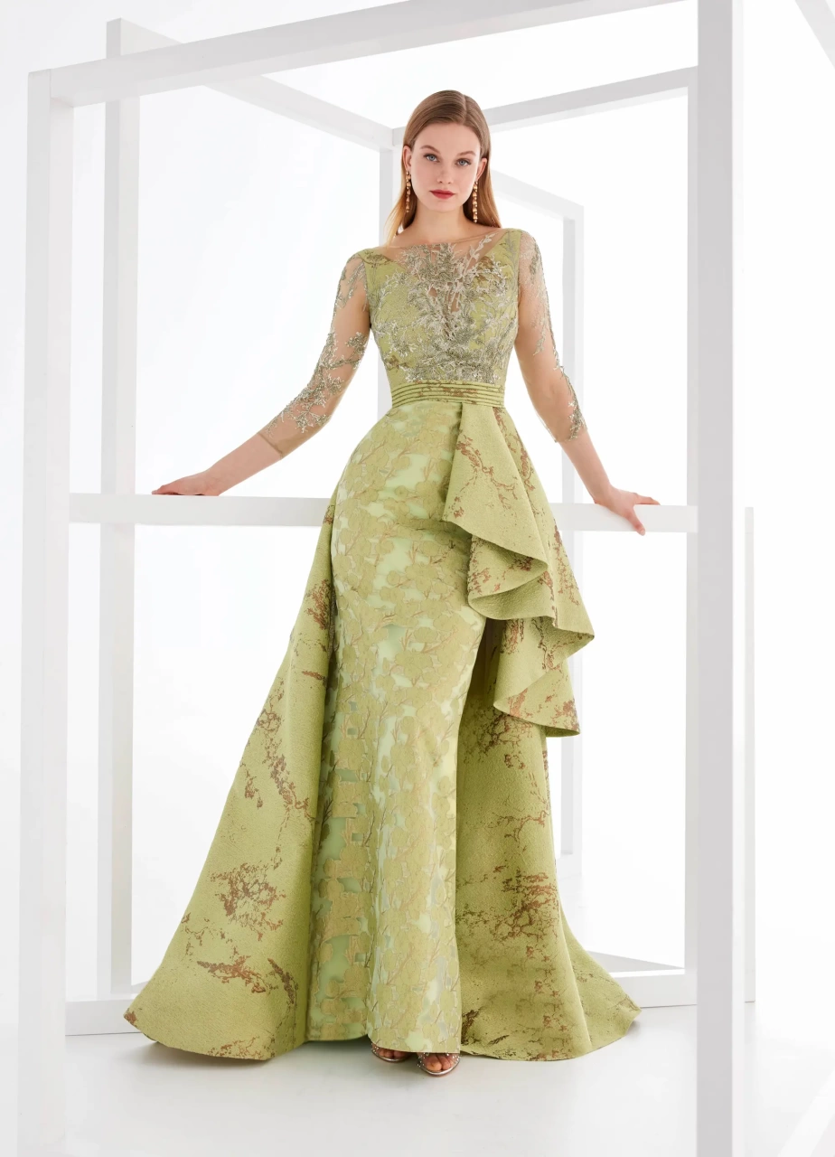 Vestito da cerimonia color verde chiaro lungo con maniche lunghe velate e pizzo ton-sur-ton, gonna lunga a sirena, sovragonna con elegante stampa a contrasto e pieghe asimmetriche sul fianco sinistro, bustino con scollo a V profondo velato - Lo Specchio Atelier Abiti da Cerimonia a Verona