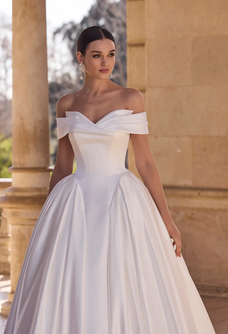 Abito da sposa semplice da principessa con vita basca, scollo off-shoulder con dettaglio particolare, gonna strutturata ampia in mikado drappeggiata sui fianchi con fiore 3D scenografico sul retro