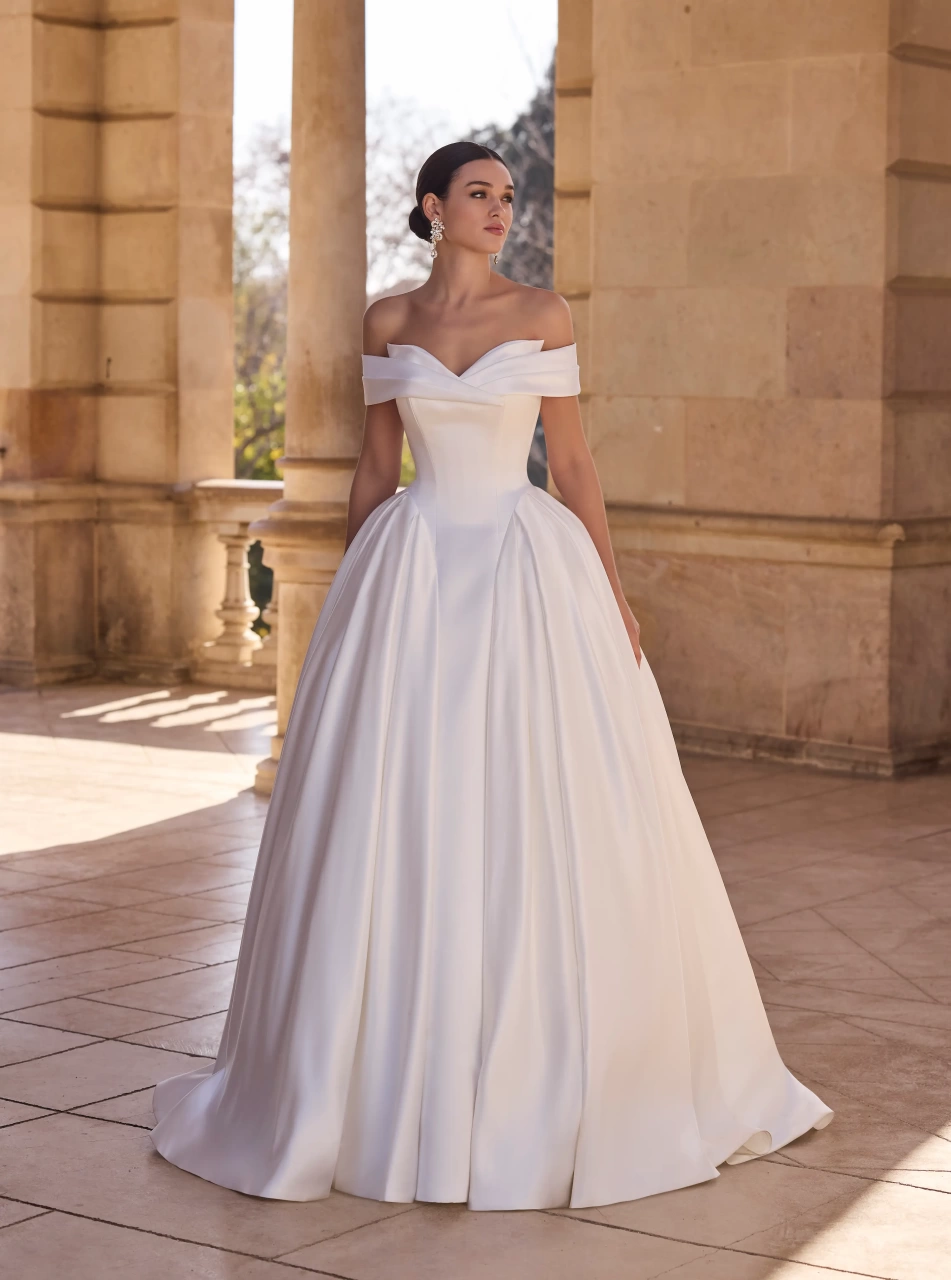 Abito da sposa semplice da principessa con vita basca, scollo off-shoulder con dettaglio particolare, gonna strutturata ampia in mikado drappeggiata sui fianchi con fiore 3D scenografico sul retro