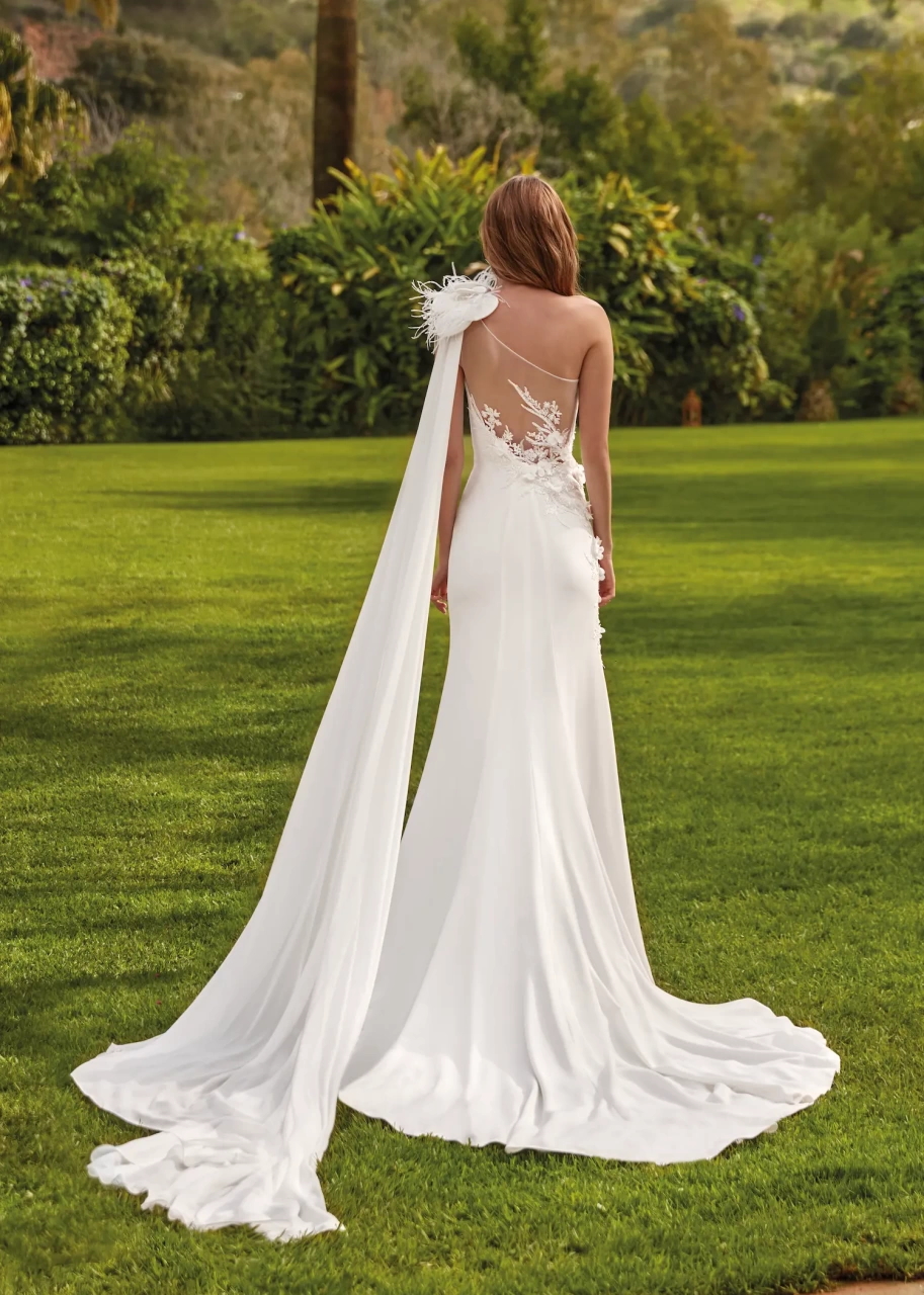 Vestito da sposa a sirena particolare con corpino monospalla drappeggiato con dettaglio di piume sulla spalla, schiena scoperta in pizzo effetto tattoo, applicazione di fiori 3D su vita e fianco destro e strascico in chiffon